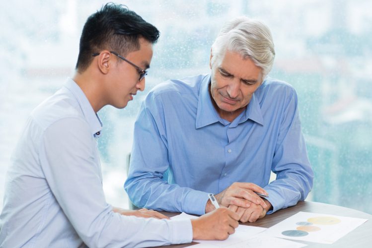 Concentrated senior businessman listening to assistant, who explaining business idea. Asian manager writing on paper. Both men sitting at table and working as team. Teamwork concept